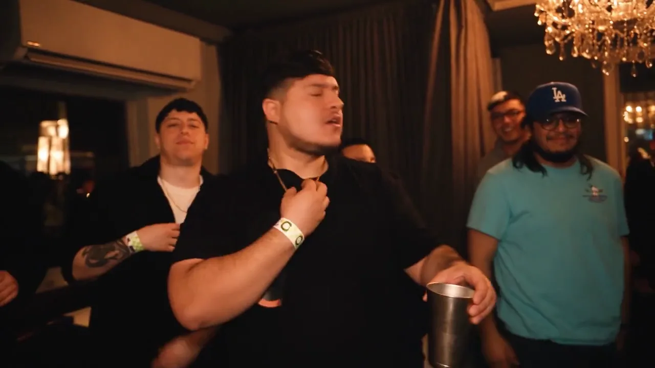 Original MV frame at twelve seconds: performer turning with a hand at his collar and a cup at waist level in a crowded room